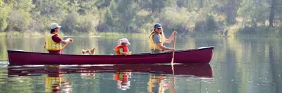 Three individuals and a dog canoeing on a serene lake surrounded by forested mountains.