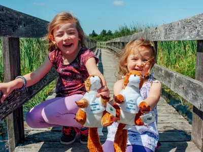 Two children smiling and sitting on a wooden bridge, each holding a stuffed toy.