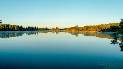 Panoramic view of a serene lake under a clear sky at sunrise, with trees reflecting on the water's surface.