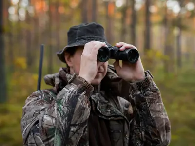 A person in camouflage clothing and a hat using binoculars in a forest.