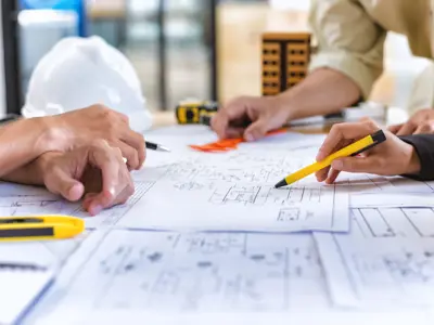 Two people examining and discussing architectural plans spread out on a table, with various tools like pencils and a measuring tape visible, in an office setting.