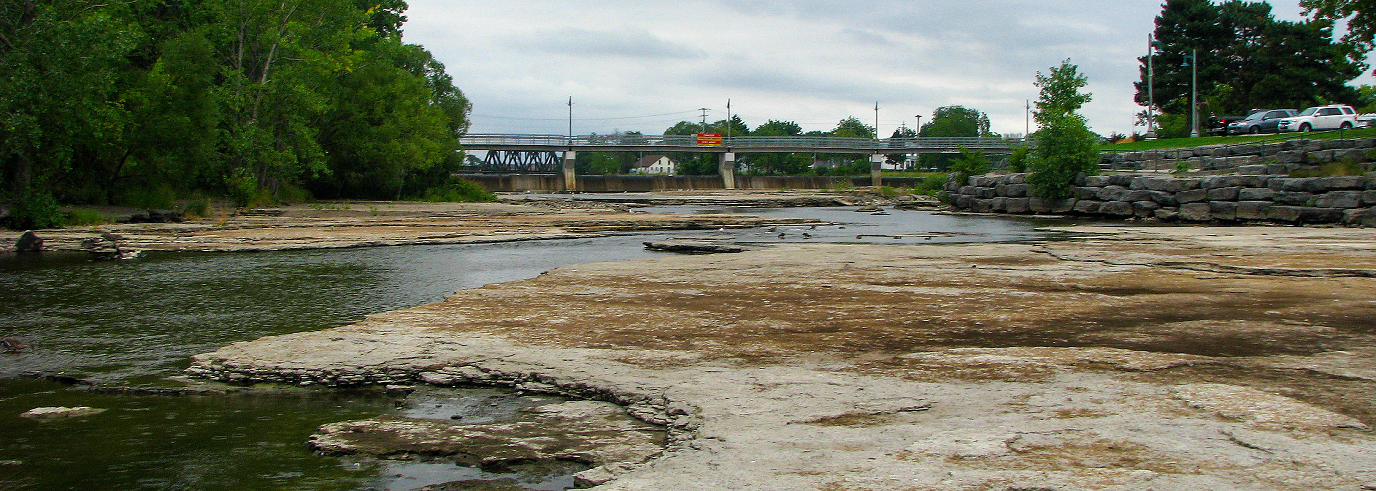 View of a shallow river with exposed rocky bed leading towards a bridge.
