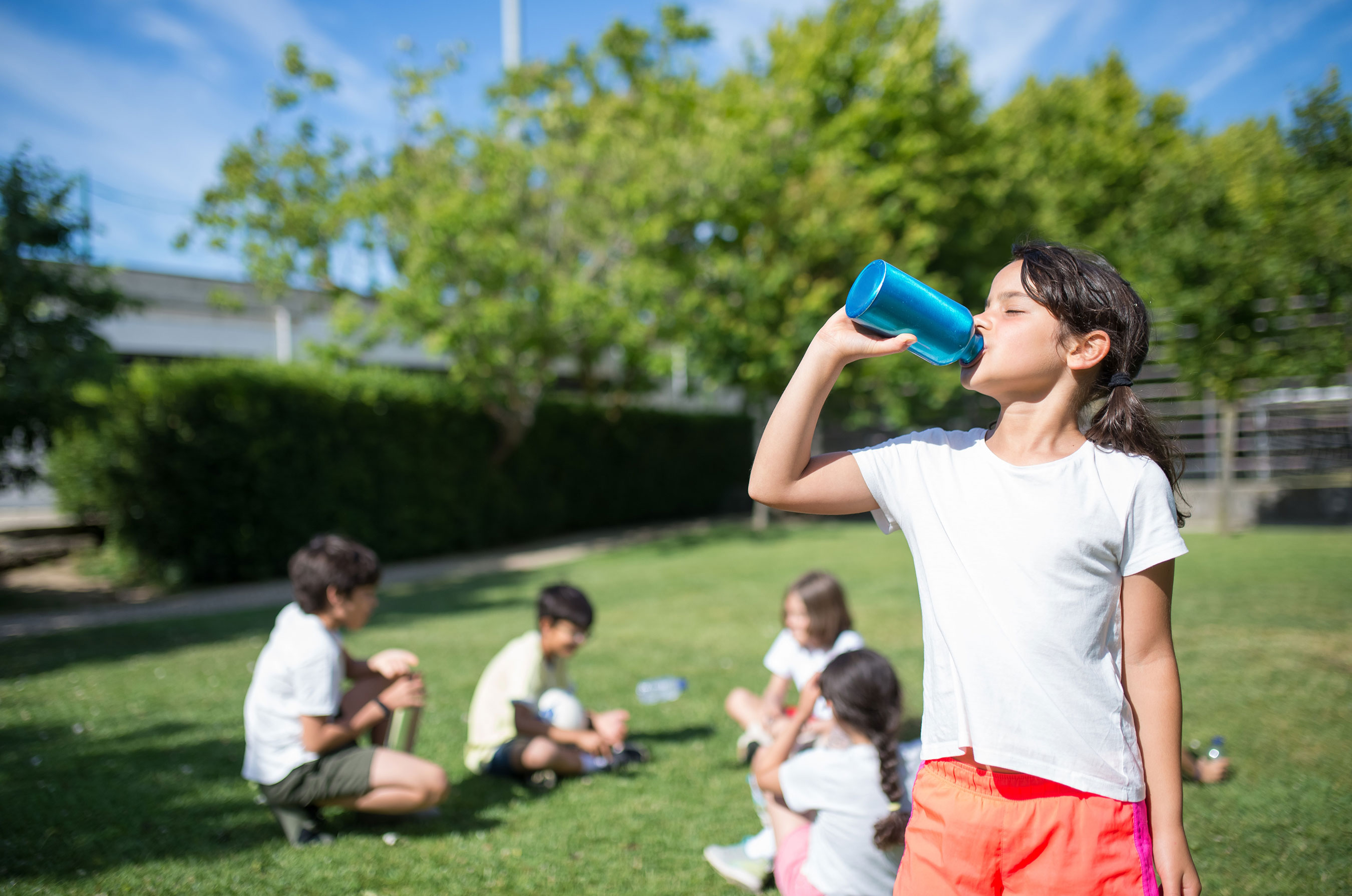 A child drinks from a blue cup in a sunny park, while other children sit on the grass in the background.