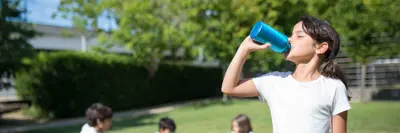 A child drinks from a blue cup in a sunny park, while other children sit on the grass in the background.