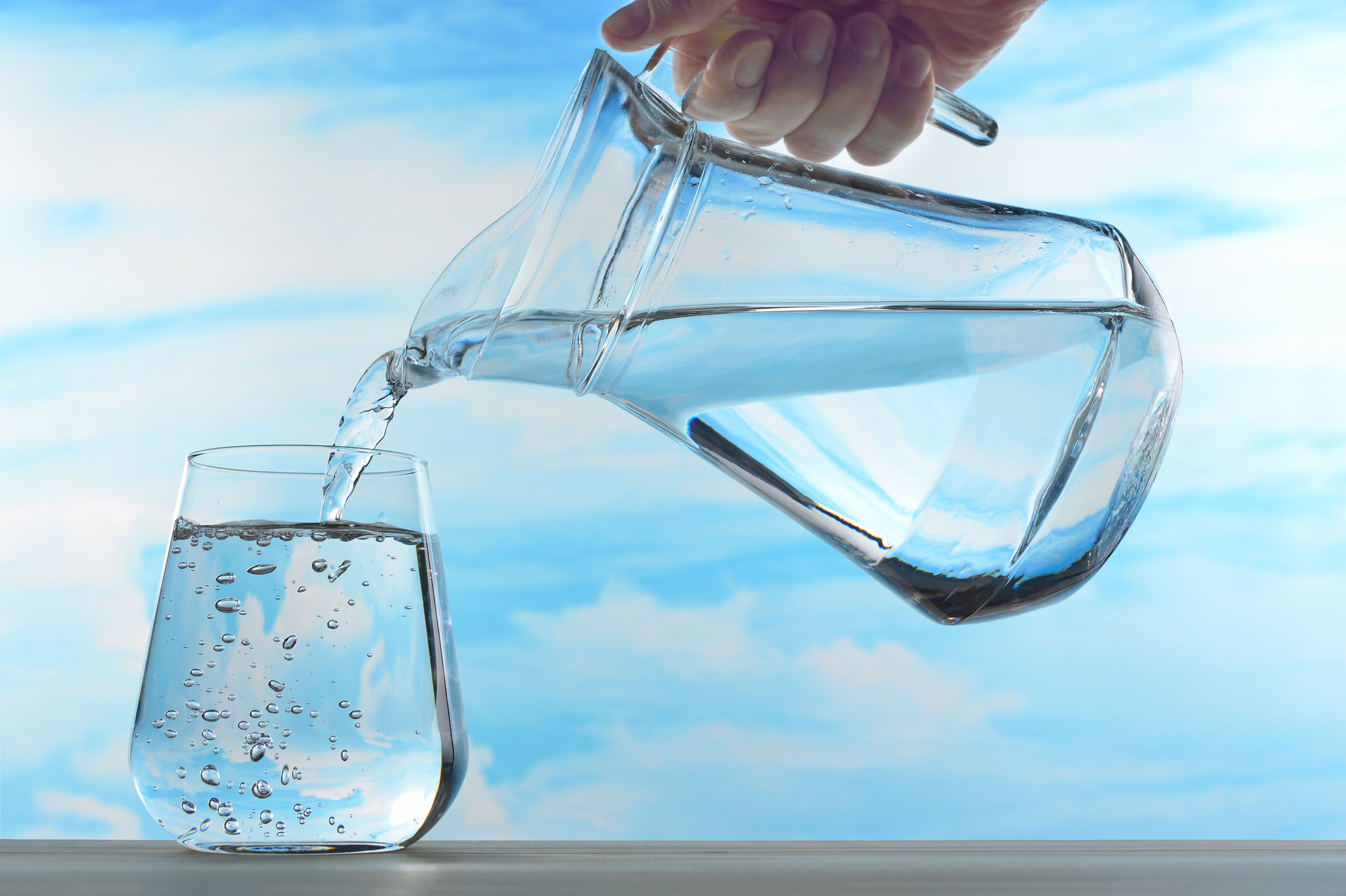 Water being poured from a clear pitcher into a glass against a sky-blue background.