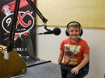 A child sits smiling at a radio station setup, wearing headphones and sitting next to a microphone, with a banner behind featuring "91X" logo.