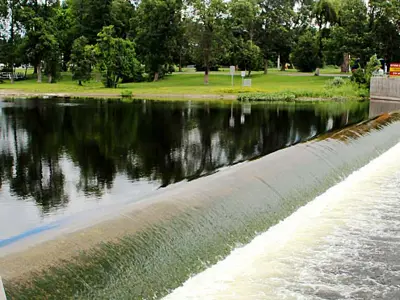 View of a river with a dam spreading across, water is flowing past a park and under a bridge, with lush greenery and a clear sky overhead.