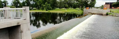 View of a river with a dam spreading across, water is flowing past a park and under a bridge, with lush greenery and a clear sky overhead.
