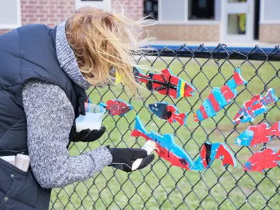 An individual hanging colorful fish cutouts on a chain-link fence outside a school building.