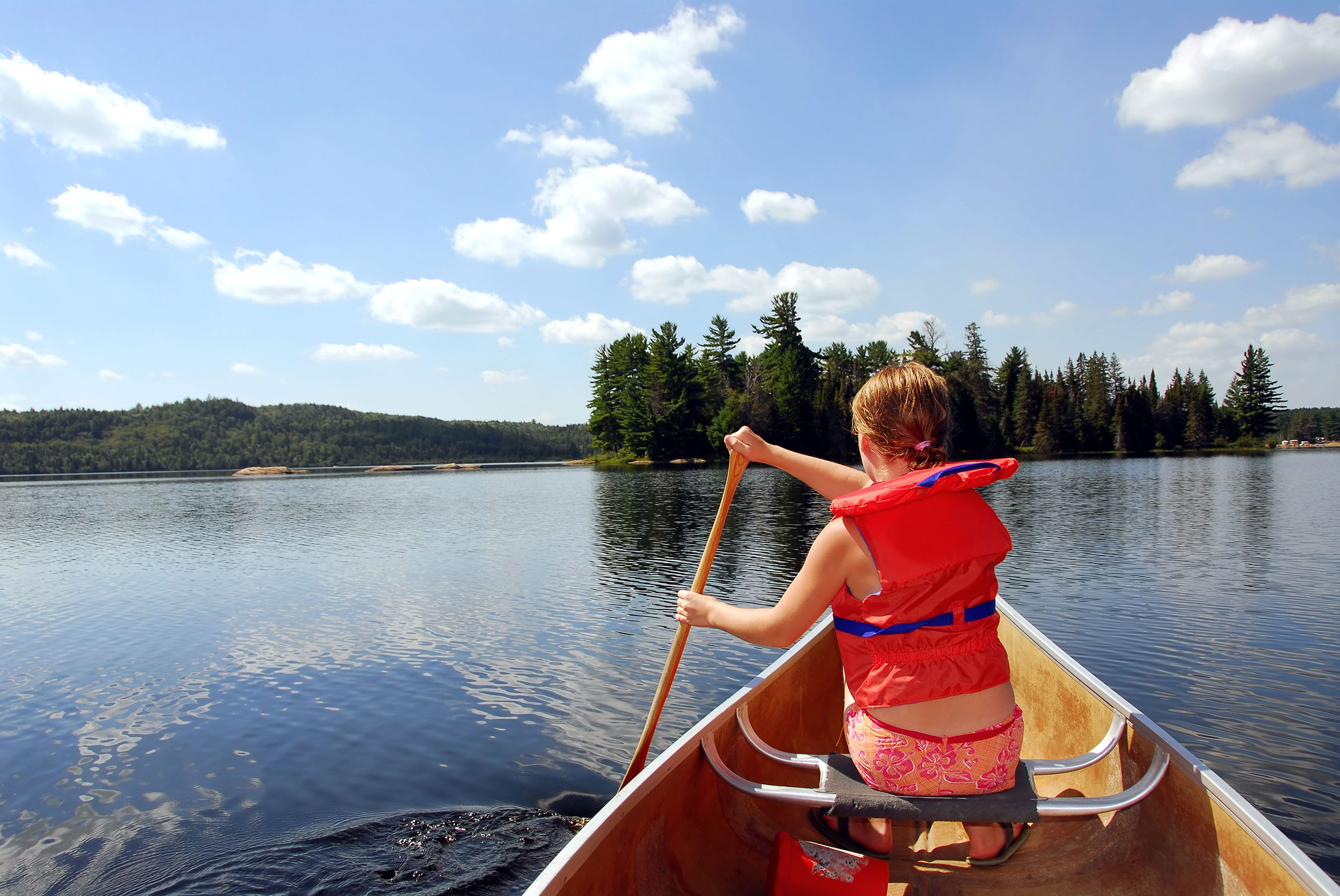 Person in a life jacket paddling a canoe on a serene lake with wooded islands in the background under a clear blue sky.