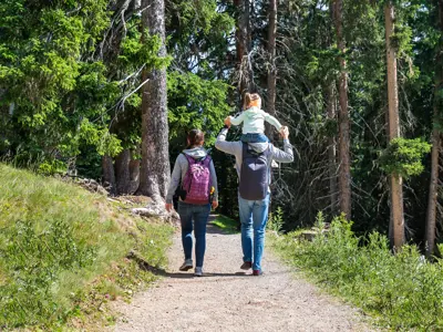 Three people hiking on a forest trail, with one individual carrying another on their shoulders.