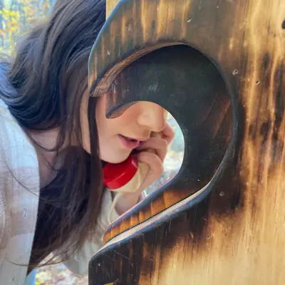 A woman in nature, stands at phone booth using a unconnected telephone 