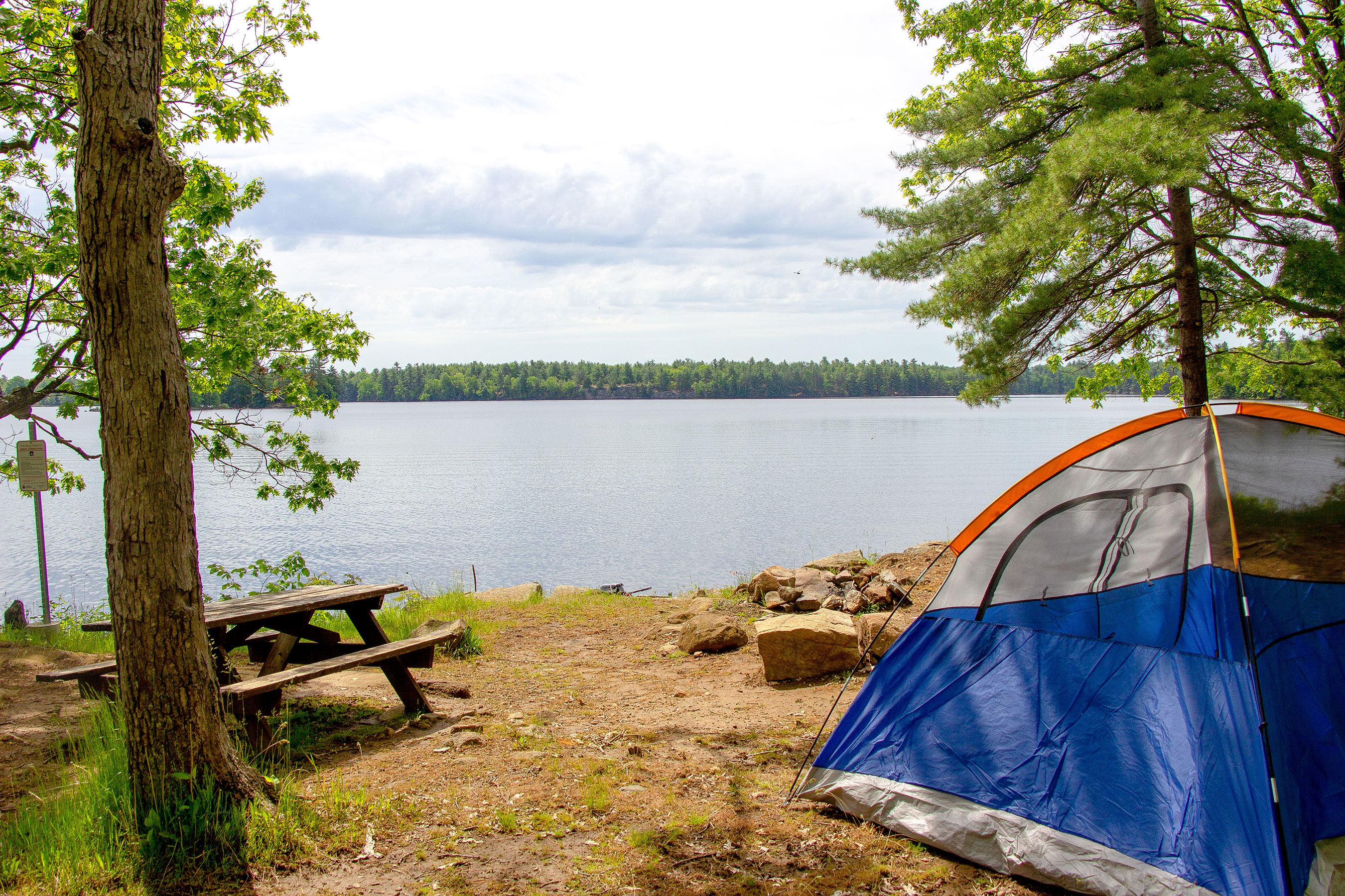 A serene camping setup beside a lake with a tent and a picnic table, surrounded by trees under a cloudy sky.