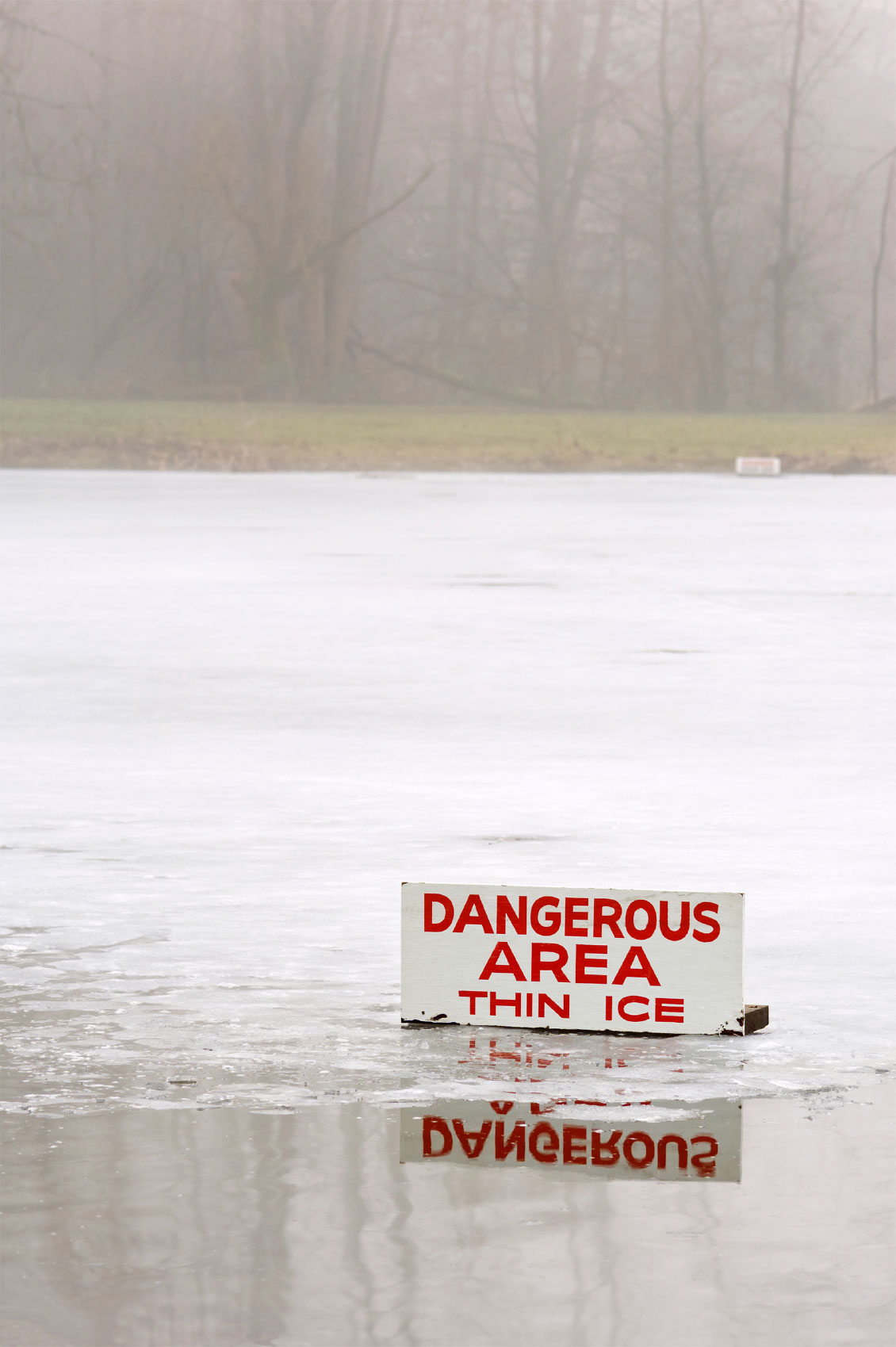 A warning sign reading "DANGEROUS AREA THIN ICE" stands partially submerged in a frozen lake with foggy, tree-lined shores in the background.