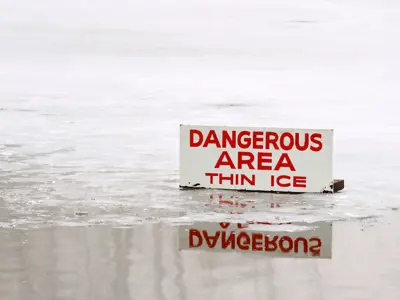 A warning sign reading "DANGEROUS AREA THIN ICE" stands partially submerged in a frozen lake with foggy, tree-lined shores in the background.