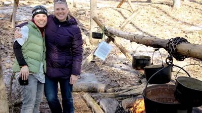 Two people smiling and standing in front of an outdoor campfire with pots hanging over the flames.