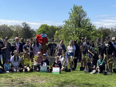 Group of people participating in a tree planting event at a park with a river in the background, accompanied by the Belleville Sens mascot.