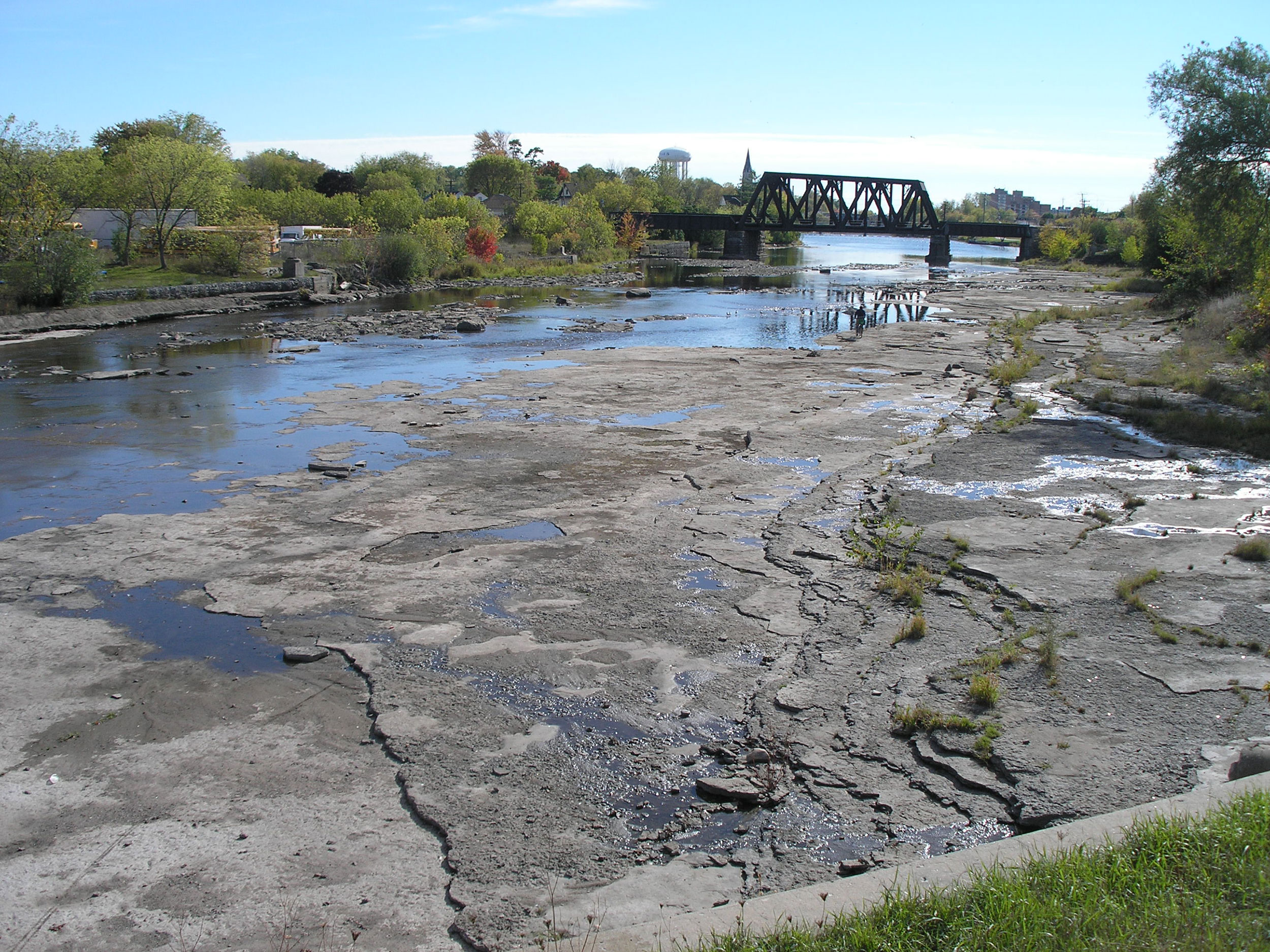 View of a shallow riverbed with patches of water. In the background, there is a metal bridge crossing the river.