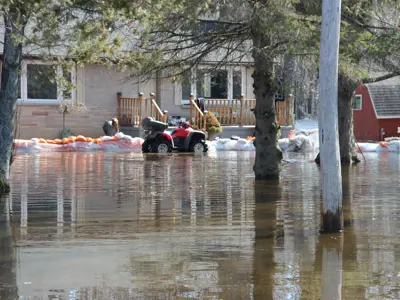 Flooded residential area. Sandbags line part of the water’s edge to mitigate flooding, and homes and trees are visible in the background.