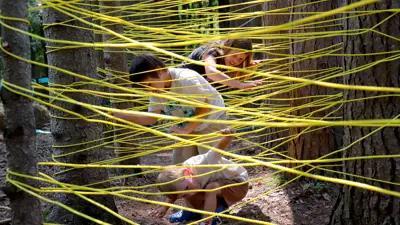 Children navigating through a yellow rope maze tied between trees in a forested area.