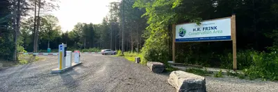 Entrance to H.R. Frink Conservation Area with a sign displaying the name, parking barriers, and a pedestrian walkway surrounded by dense trees under a clear sky.