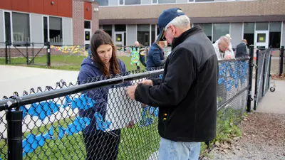 Two individuals attaching painted, wooden fish to a chain-link fence outside a school building, with other people in the background participating in the activity.