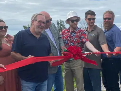 Group of individuals participating in a ribbon-cutting ceremony outdoors, with a logo that includes a tree and two hills visible on a sign next to them.