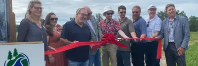 Group of individuals participating in a ribbon-cutting ceremony outdoors, with a logo that includes a tree and two hills visible on a sign next to them.