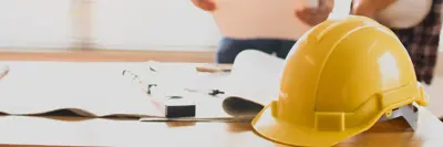 Two individuals examining blueprints at a work desk with various tools and a yellow hard hat visible in the foreground.