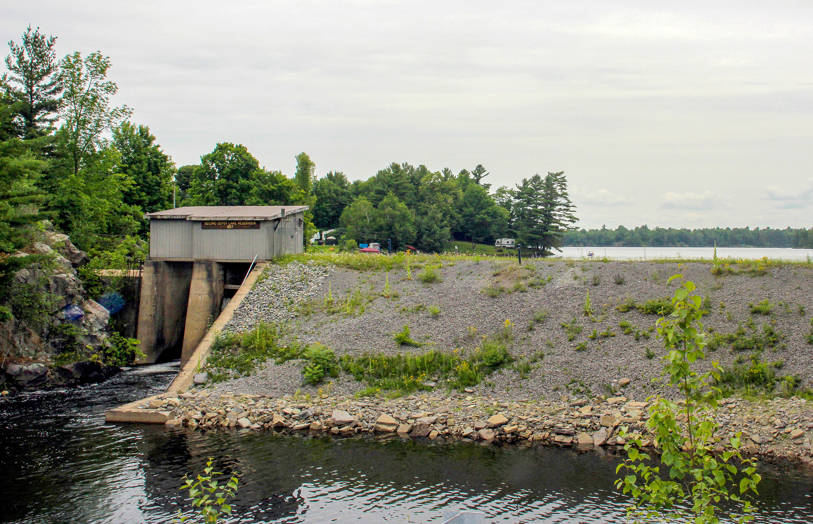 A scenic view of a covered bridge over a small river, with a forested area in the background and a cloudy sky.